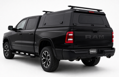 A black Ram pickup truck with a black hard canopy cover on the bed, shown from a rear three-quarter angle on a white studio background. The truck features a roof rack, black wheels, and a visible 'IRONMAUL' license plate frame on the rear bumper.