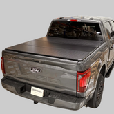 Black Ironhaul hard tri-fold tonneau cover installed on a gray pickup truck with a light neutral background